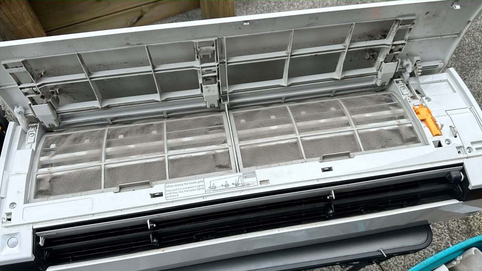Indoor heat pump opened up, showing filters and intake grille clogged with grey dust before cleaning